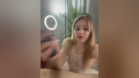 Video of a young blonde woman with fair skin, wearing a white lace top, sitting in front of a lighted ring light, with ferns in the background.