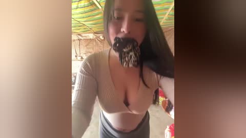 Media: Video of an Asian woman with long black hair, wearing a low-cut, beige ribbed top revealing cleavage, eating a black and white striped lollipop under a green and yellow striped canopy in a rustic setting.