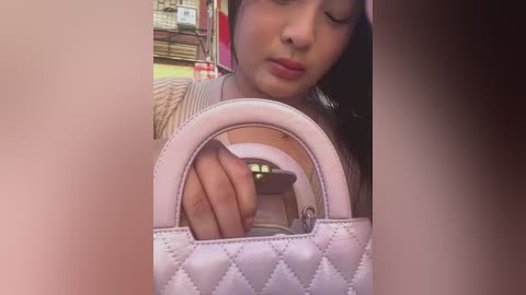 Video of a young woman with medium skin tone, Asian ethnicity, wearing a pink headband, a beige sweater, and a white quilted handbag. She is holding the bag with a closed right hand. Background shows a blurred indoor setting with colorful items.