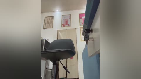 Video of a narrow, dimly-lit hallway with a black plastic chair on the left, beige walls adorned with framed art, and a closed door on the right.