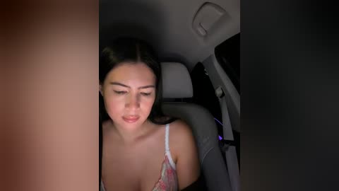 Video of a young Asian woman with long black hair, wearing a floral-patterned bra, seated in a car at night, looking down, illuminated by car lights.