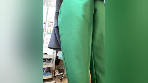 Video of a person in green scrubs standing in a hospital hallway. The background shows a white wall, medical equipment, and a door with signs. The image is taken from a low angle, emphasizing the person's legs and feet.