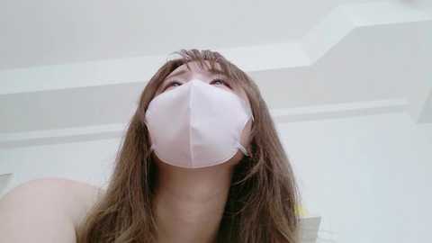 A video of a young woman with long brown hair, wearing a white face mask, taken from a low angle, showing her upturned face against a white ceiling.