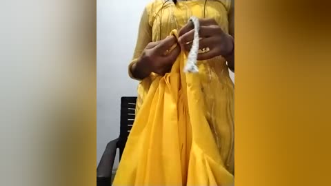 Video of a person in a yellow saree, holding a white thread, standing against a plain white wall. Background includes a black plastic chair.