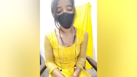 Video of a woman in a yellow saree and mask, sitting in a chair, with a yellow curtain behind her.