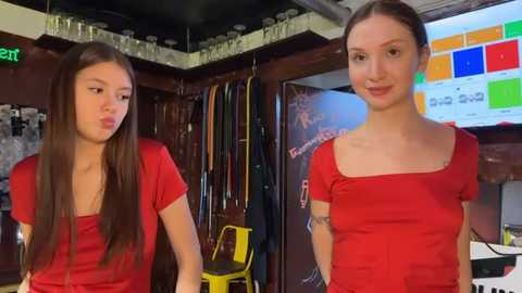 Video of two young women with long brown hair in red shirts, standing in a cluttered, dimly lit room with a chaotic background of hanging clothes, chairs, and a colorful poster.