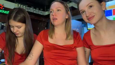 Video of three young women in red tops, with one wearing a tattoo on her arm, in a dimly lit bar setting.