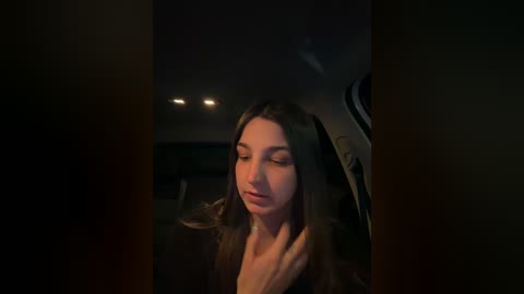 Video of a young woman with long brown hair, fair skin, and closed eyes, wearing a brown coat, seated in a dimly lit car at night, looking serene.