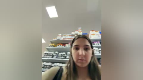 A video of a young woman with long brown hair, wearing a beige sweater, standing in a pharmacy with shelves filled with various medicines. The image is slightly blurred.