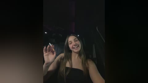 Video of a smiling young woman with long brown hair, wearing a strapless black dress, holding a lit cigarette, against a dimly lit background with blurred shapes and textures.