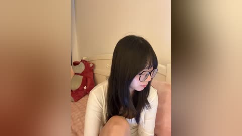 Video of a young Asian woman with straight black hair, wearing glasses and a white top, sitting on a bed with a red pillow and beige blanket, in a dimly lit room.