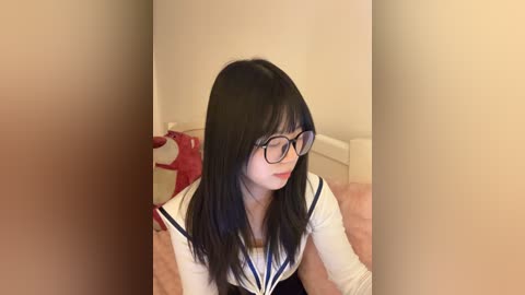 Video of an Asian woman with long black hair, wearing glasses and a Japanese schoolgirl uniform, sitting on a bed with beige sheets.