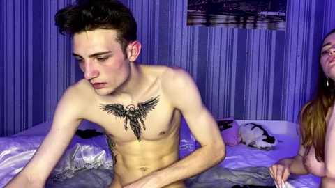 Video of a young, shirtless, lean Caucasian man with short dark hair, a detailed chest tattoo, and a woman with long brown hair, partially visible, in a dimly lit room with a purple hue.