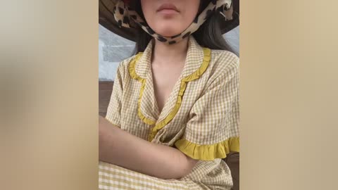 Video of a young woman with fair skin, wearing a yellow and white checkered dress with a frilled neckline, and a cow-print mask. Background shows a wooden surface and a white bedsheet.