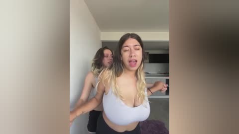 Video of two young women in a hallway. One, in a white crop top, has long, blonde hair and is startled. The other, with long brown hair, is behind her, looking concerned.