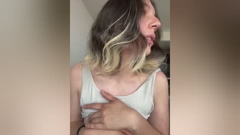 Video of a young woman with light skin and shoulder-length, wavy, blonde-streaked brown hair, wearing a white tank top. She has a hand on her chest, tongue sticking out in a playful, exaggerated manner. Background is a neutral, indoor setting with soft lighting.