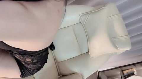 Video of a woman's upper body in black lace lingerie, sitting on a cream-colored leather armchair. She has pale skin and is indoors, with a white curtain and a blurred bookshelf in the background.