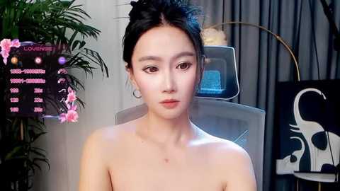 Video of a fair-skinned Asian woman with dark hair, wearing minimal makeup, sitting topless in a modern room with a potted plant, digital screen, and black curtains in the background.