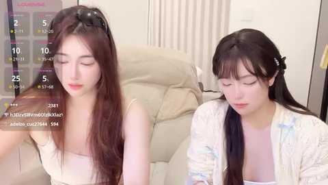 A video featuring two young East Asian women with long black hair, one in a white floral cardigan, the other in a white top, seated on a beige couch in a living room.