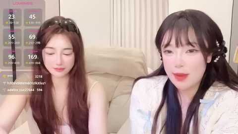 Video of two East Asian women with long black hair, one wearing a headband, sitting on a beige couch, in a light-toned room.