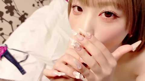 Video of an Asian woman with light skin, brown hair, and long manicured nails, wearing a white robe, with a pink vibrator and black hair accessories on a floral-patterned background.