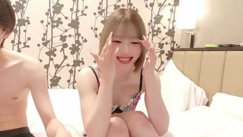 Video of a smiling, young Asian woman with straight, shoulder-length blonde hair, wearing a floral bra, sitting on a bed in a modern bedroom with white sheets and a patterned wall.