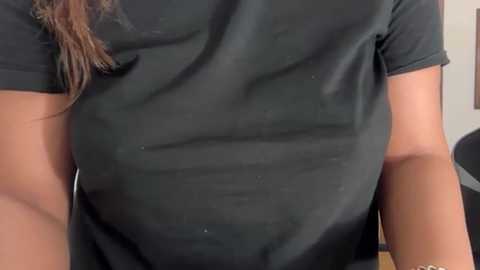 Video of a person's torso wearing a black T-shirt, with a glimpse of brown hair visible. The background is blurred, showing a plain wall. The person appears to be seated, with the shirt's texture and slight creases visible.