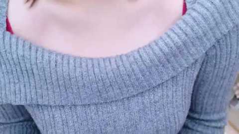 A close-up video of a woman's upper torso in a light gray, ribbed-knit sweater with a round neckline, showcasing her fair skin and a hint of a red bra strap.