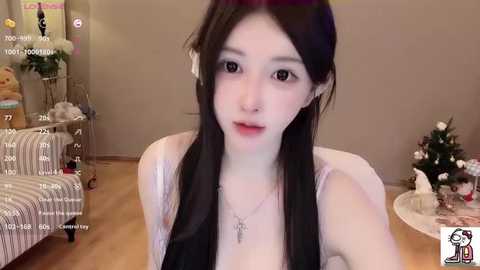 Video of a young East Asian woman with pale skin, long black hair, and a slender physique. She wears a white tank top, and her expression is neutral. Background features a beige wall, a Christmas tree, and a striped sofa.