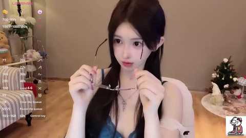 Video of an East Asian woman with long black hair, small breasts, wearing a low-cut blue dress, adjusting her glasses in a cozy, Christmas-decorated living room.