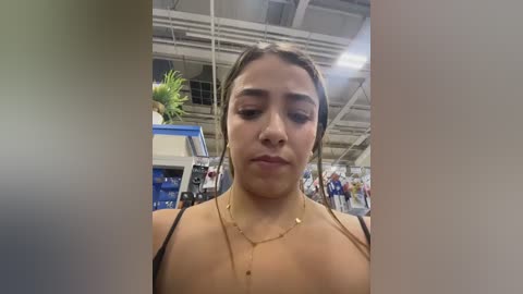 Video of a young woman with medium skin tone, straight brown hair, and a neutral expression, wearing a tan top and gold necklace. She stands indoors at an event, with a palm tree and exhibition booths in the background.