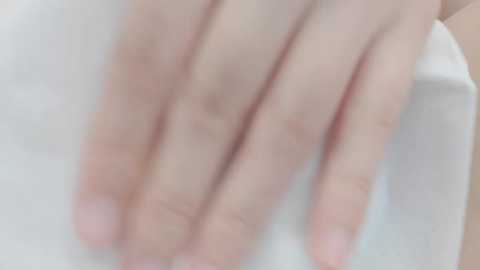 Close-up video of a fair-skinned hand with well-manicured nails resting on a white, smooth surface, likely a table or counter. The background is blurred, focusing attention on the hand and its delicate touch.