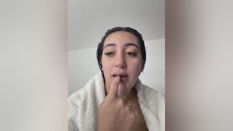 Video of a young woman with medium brown skin and dark hair, wearing a white towel and biting her finger, in a minimalist white room.