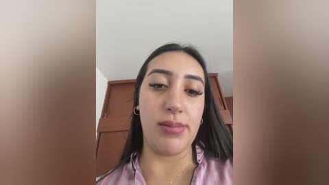 A video of a young woman with medium skin tone, long black hair, and full lips, wearing a pink tie-dye top, against a brown wooden background.