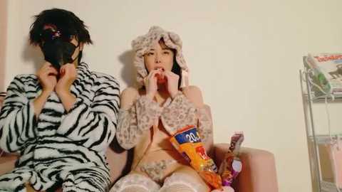 Video of two Asian women in zebra-pattern pajamas eating ice cream cones and drinking soda, sitting on a couch against a beige wall, with a shopping cart in the background.