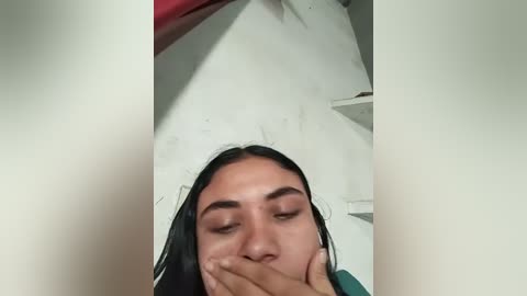 A close-up video of a young woman with long black hair, dark eyes, and fair skin, covering her mouth with her hand, set against a plain white wall background.