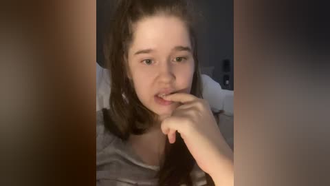 Video of a young woman with light skin, brown hair, and green eyes, biting her index finger, wearing a grey shirt, in a dimly-lit bedroom.