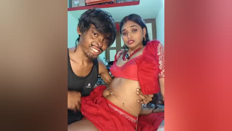 Video of a young, dark-skinned man in a black tank top and a pregnant woman in a red sari, smiling, in a brightly lit room.