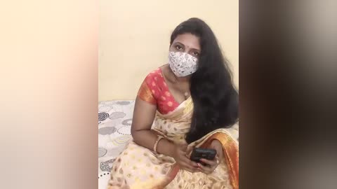 Video of a South Asian woman with long black hair, wearing a pink and gold sari, face mask, and bracelet, seated on a bed with white and grey patterned sheets.
