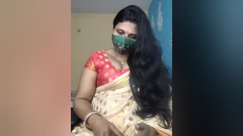 Video of a South Asian woman with long black hair, wearing a red saree with gold accents, a green mask, and a gold bracelet, sitting indoors against a beige and blue wall.