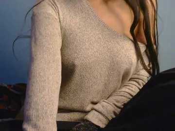 Video of a woman with light skin, wearing a beige knitted sweater, revealing her medium-sized breasts, sitting on a black couch, with a blue wall background.