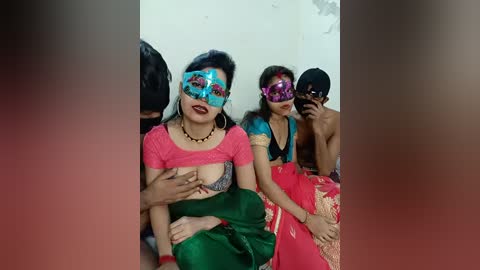 Video of four women in traditional Indian attire with colorful masks, one breastfeeding, sitting on a bed in a simple room.