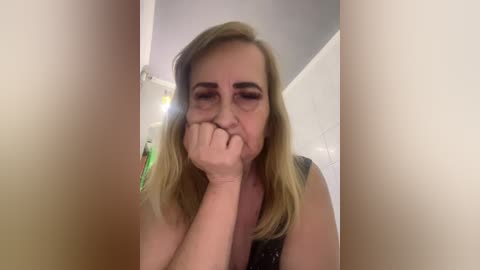 Video of a middle-aged, fair-skinned woman with shoulder-length blonde hair, wearing a sleeveless black top, covering her mouth with her hand, looking upset. Background shows a white wall and a green object.