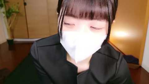 Video of an Asian woman with straight black hair and bangs, wearing a black coat and white mask, in a dimly lit indoor room with a potted plant and wooden floor.