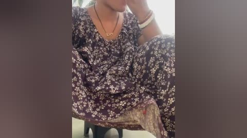 Video of a person sitting with a purple floral dress, wearing a gold necklace, and bracelets. Background is blurry.