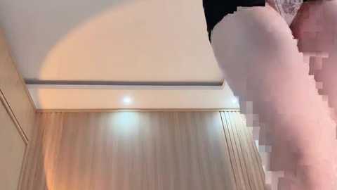 A video captures a woman's legs in white stockings, positioned in a modern, beige-toned room with a ceiling light. The image is slightly blurred and pixelated, adding a surreal, dreamlike quality.