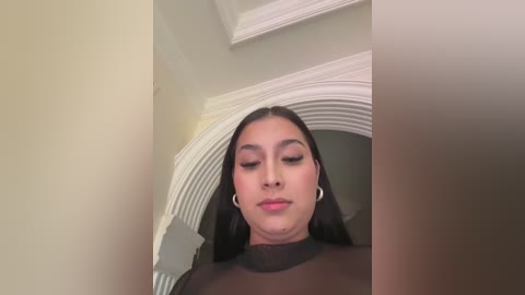 Video of a young woman with long black hair, wearing large hoop earrings, standing in a doorway with an arched white frame, smiling gently.