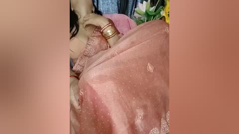 Video of a woman in a pink, embroidered sari with gold bangles, lying on a bed with a blue sheet, partially covered by a blanket. The background includes green plants and a yellow flower.