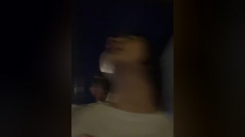 A blurry video of a person with a dark complexion, possibly African American, wearing a white shirt, mouth open, captured in a dimly lit room with a dark background.