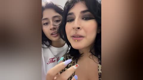 Video of two young women with medium brown skin, dark hair, and dark makeup, one with a nose ring, wearing leopard print tops, standing close together.